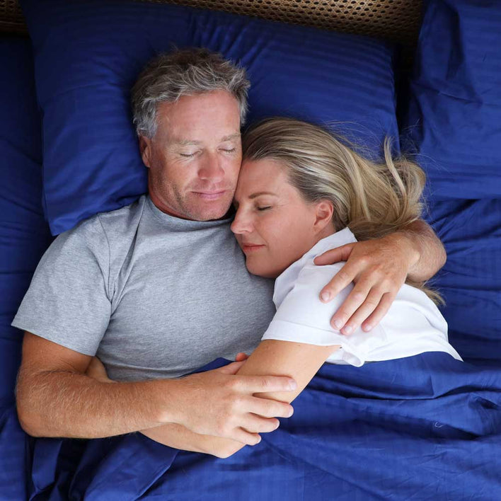 A man and women cuddling in a soft bed complimented by navy bed sheets.
