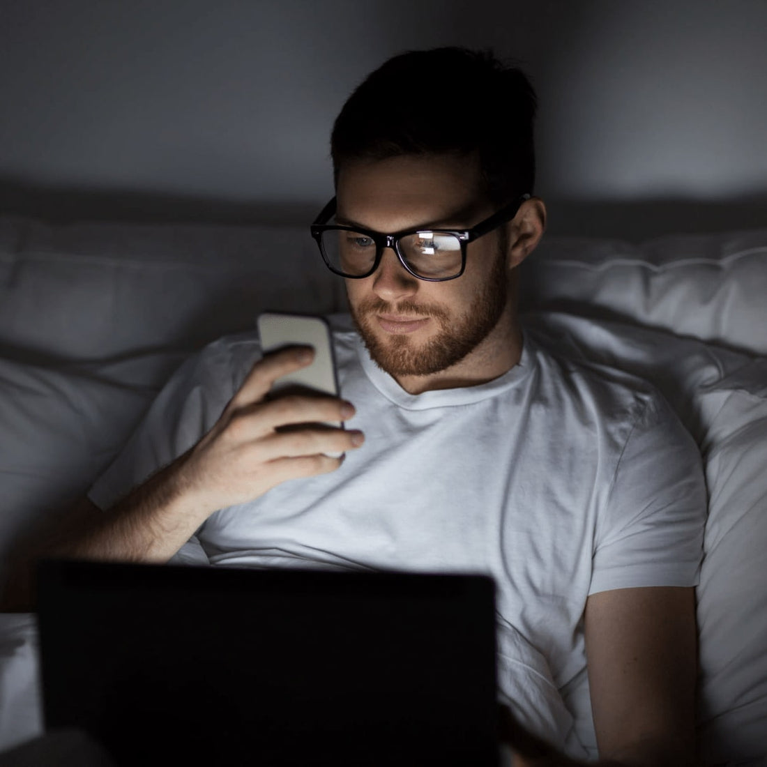 Young man lying down on his bed at night with blue light glasses and using his phone and laptop.