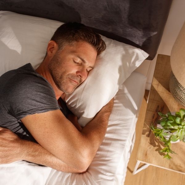 Middle aged man cuddling his pillow while asleep on his mattress.