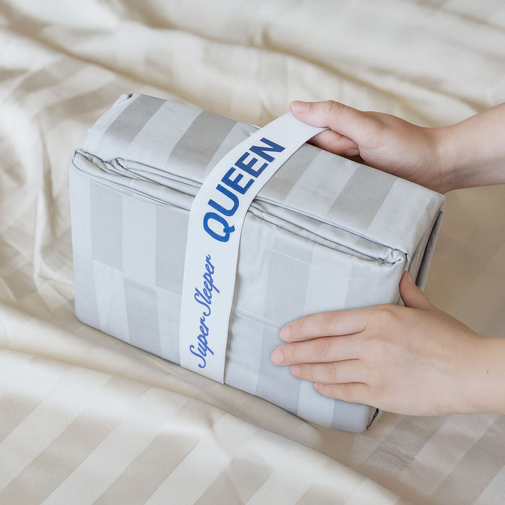 A person holds a folded, light grey, striped Super Sleeper Pro TEST broken variant os bedding set labeled QUEEN on a beige striped bedspread, demonstrating an ideal linen cupboard organiser sheet storage solution.