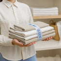 A person in a light shirt holds a stack of QUEEN beige and white sheets, neatly secured with Super Sleeper Pro® Bed Sheet Organiser Bands. Shelves with more linens and a woven basket can be seen in the background.