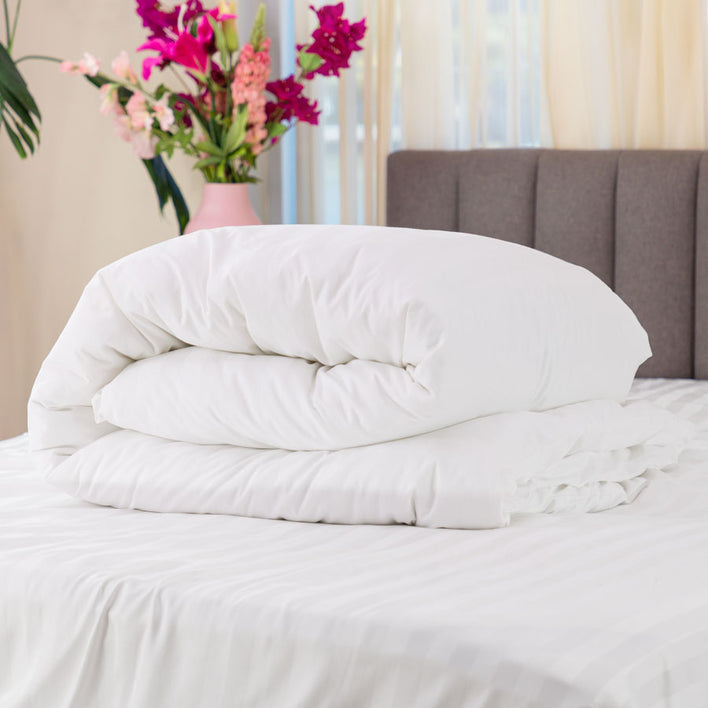 A neatly folded Royal Deluxe Hotel Quality 100% Cotton Quilt Cover by Super Sleeper Pro rests on a bed with a white striped sheet. A vase filled with pink and red flowers sits near a window draped in sheer curtains.