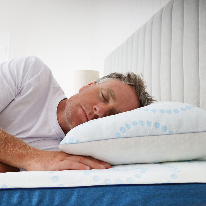 A middle-aged man with gray hair sleeps on his side, resting on a white pillow with blue circles. He enjoys extra comfort from the Super Sleeper Pro #1 Customer Choice Straight 2 Sleep® Mattress Topper with Cooling Technology and bonus free pillows.