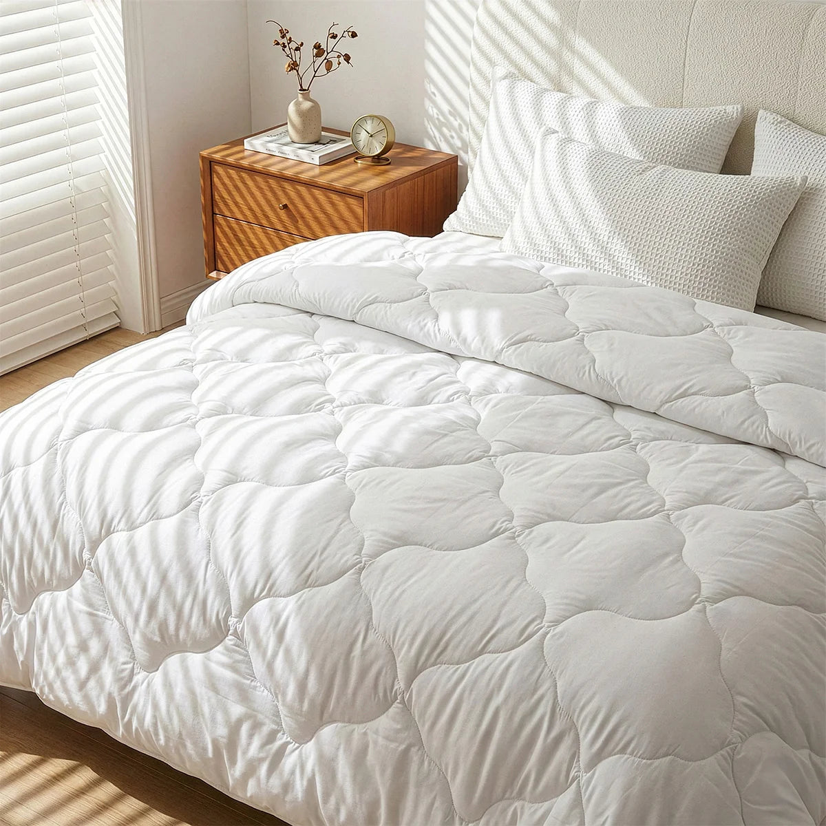 A bed features the Super Sleeper Pro Quilt 365™—two plush quilts that snap together—plus two FREE luxury pillows. A wooden nightstand holds a vase and round clock, basking in sunlight through window blinds.