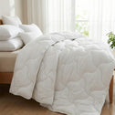 A bed features the Super Sleeper Pro Quilt 365™—two plush quilts that snap together for cosy warmth—plus two free luxury pillows. Sunlight fills the room, with a green plant resting on the floor nearby.