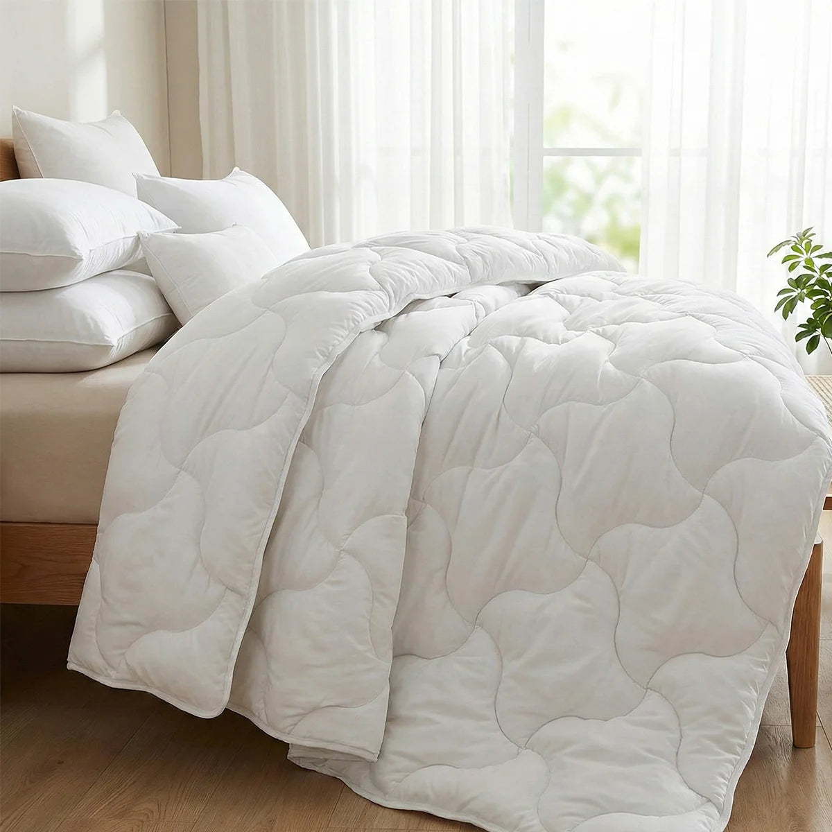 A bed features the Super Sleeper Pro Quilt 365™—two plush quilts that snap together for cosy warmth—plus two free luxury pillows. Sunlight fills the room, with a green plant resting on the floor nearby.