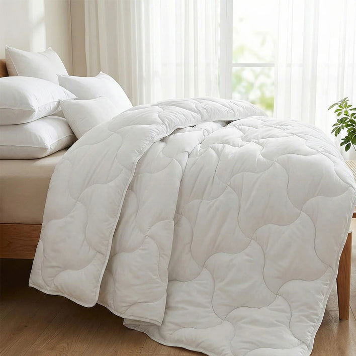 A bed features the Super Sleeper Pro Quilt 365 All Season 2-in-1 Adjustable Quilt Set with 2 free Luxurious Marshmellow Pillows, placed by a bright window with sheer curtains and a small green plant for a fresh touch.