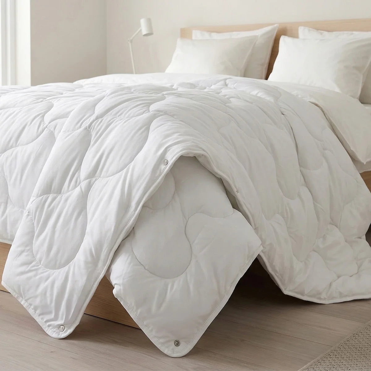 A bright, minimal bedroom features the Super Sleeper Pro Quilt 365™—two plush quilts that snap together for cozy warmth—draped on a bed with white luxury pillows, a beige headboard, light floors, and a white lamp in the background.