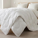 A bright, minimal bedroom features the Super Sleeper Pro Quilt 365™—two plush quilts that snap together for cozy warmth—draped on a bed with white luxury pillows, a beige headboard, light floors, and a white lamp in the background.