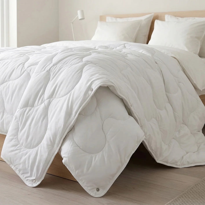 A bright, minimal bedroom features the Super Sleeper Pro Quilt 365™—two plush quilts that snap together for cozy warmth—draped on a bed with white luxury pillows, a beige headboard, light floors, and a white lamp in the background.