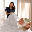A woman in navy pajamas puts a white Super Sleeper Pro Quilt 365™ All Seasons Two Plush Quilts on a bed. An inset shows her fastening a snap button on the cover, with light streaming through sheer curtains behind.