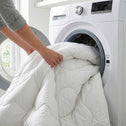 A person loads a large white Super Sleeper Pro Quilt 365™ All Seasons Two Plush Quilts into a front-loading washing machine in a bright laundry room, the door open and their sleeves rolled up.