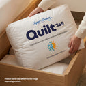 A person places a white Super Sleeper Pro Quilt 365™ All Seasons Two Plush Quilts pillow—featuring climate control icons and all-season branding—into a wooden drawer. Product name and a note about variation are visible on the pillow.