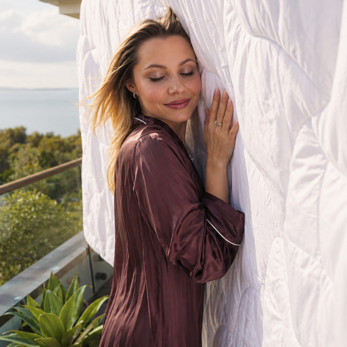 A woman in a satin robe stands on a balcony, eyes closed as she gently touches the Super Sleeper Pro Quilt 365™ All Seasons Two Plush Quilts hanging vertically, surrounded by lush greenery with water and trees in the peaceful background.