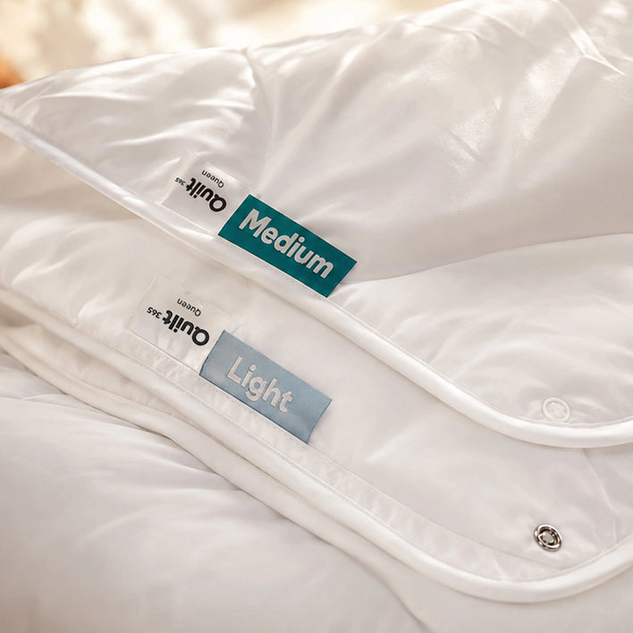 Two Super Sleeper Pro Quilt 365™ All Seasons quilts are stacked, showing a green Medium and blue Light label. Both offer clean, soft, and adjustable warmth for comfort all year round.
