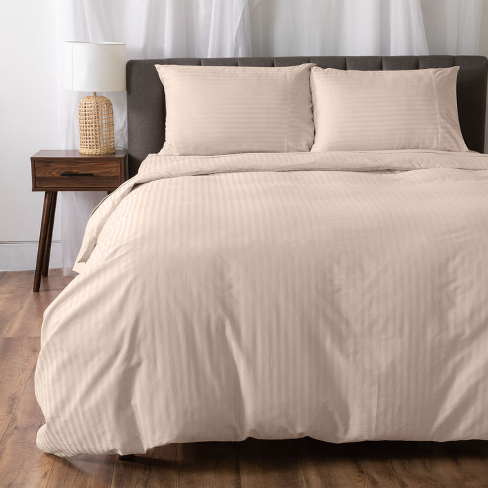 A neatly made bed features the Super Sleeper Pro Royal Deluxe Hotel Quality 100% Cotton Quilt Cover in beige stripes and two pillows against a gray upholstered headboard. A wooden nightstand with lamp stands on the wooden floor, white curtains behind.