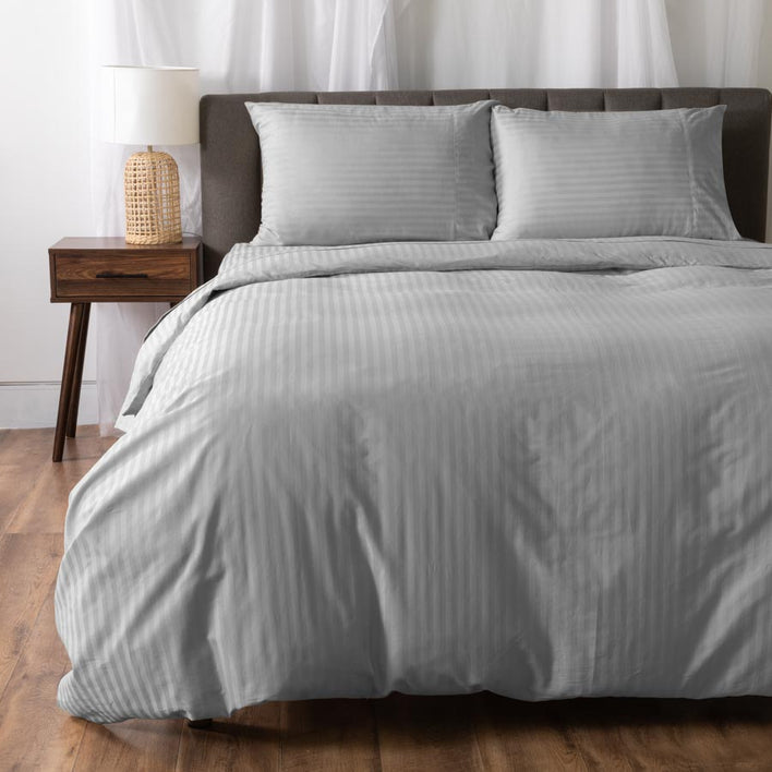 A neatly made bed features the Super Sleeper Pro Royal Deluxe Hotel Quality 100% Cotton Quilt Cover in light gray stripes with two matching pillows and a dark headboard. A wooden nightstand with a lamp stands on the wood floor, white curtains behind.