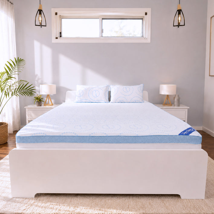 A neatly made bed with a white frame, topped with two pillows and a blue Super Sleeper Pro Straight 2 Sleep® Mattress Topper, sits in a bright bedroom. Matching nightstands with lamps flank the bed, and a plant is placed by the window adorned with white curtains.