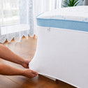 A person is pulling a white Super Sleeper Pro mattress protector over the edge of a bed with a blue and white mattress, possibly preparing to add their Straight 2 Sleep® Mattress Topper by Super Sleeper Pro, in a room featuring wood flooring and sheer lace curtains.