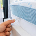 A hand is unzipping the cover of a blue and white Straight 2 Sleep® Mattress Topper by Super Sleeper Pro. The background features a window with sheer curtains and greenery visible outside.