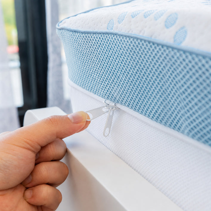 A hand is unzipping the cover of a blue and white Straight 2 Sleep® Mattress Topper by Super Sleeper Pro. The background features a window with sheer curtains and greenery visible outside.