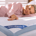 A woman lies on a bed, resting her head on a pillow and touching the Super Sleeper Pro Straight 2 Sleep® Mattress Topper with her hand, while sunlight filters through shutters in the background.