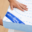 A hand presses on the corner of a Super Sleeper Pro mattress labeled “Super Sleeper Pro.” The mattress, featuring a white and blue cover with a mesh blue border and a visible brand tag on the side, is perfectly complemented by the plush comfort of the Straight 2 Sleep® Mattress Topper from Super Sleeper Pro. The entire setup rests elegantly on a wooden floor.