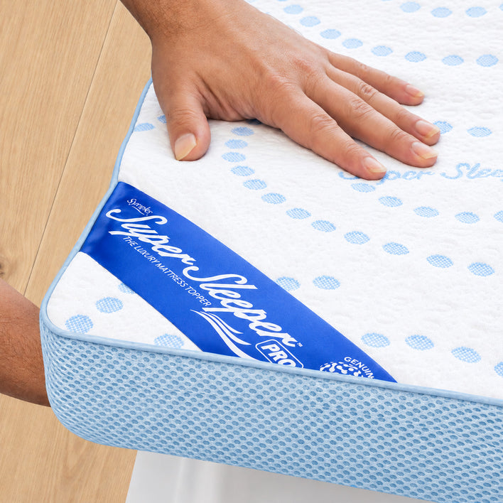 A hand presses on the corner of a Super Sleeper Pro mattress labeled “Super Sleeper Pro.” The mattress, featuring a white and blue cover with a mesh blue border and a visible brand tag on the side, is perfectly complemented by the plush comfort of the Straight 2 Sleep® Mattress Topper from Super Sleeper Pro. The entire setup rests elegantly on a wooden floor.