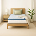 A single bed with a wooden frame features the Super Sleeper Pro [King Single] Straight 2 Sleep® Mattress Topper with Cooling Technology and free pillows. A beige blanket is draped over one corner, beside a wooden nightstand with a lamp and plant.