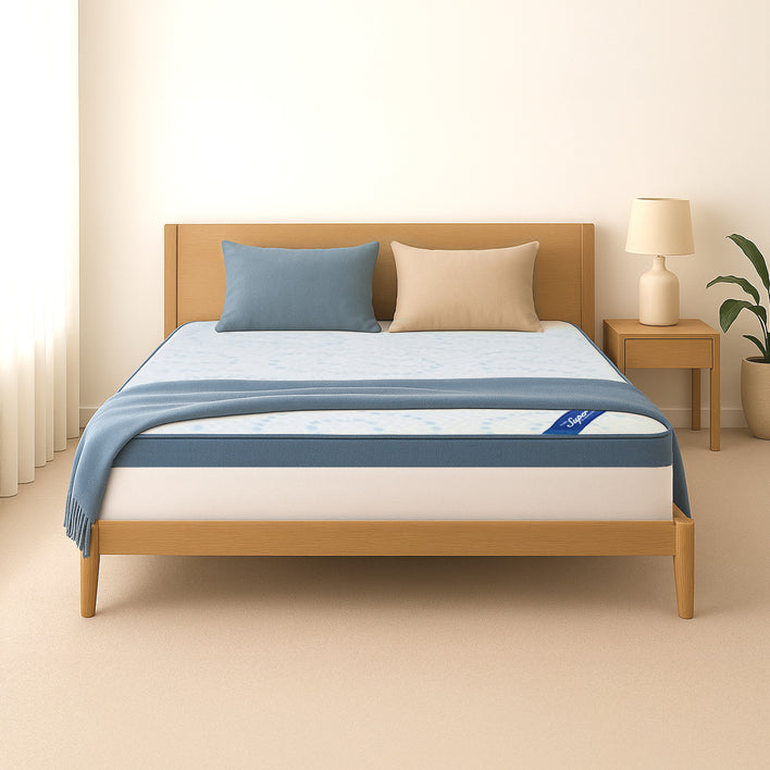 A minimalist bedroom with a light wood bed frame, blue and beige pillows, blue blanket, and white mattress topped by the Super Sleeper Pro [Double] Straight 2 Sleep® Mattress Topper with Cooling Technology + FREE Pillows. Lamp and plant on nightstand.
