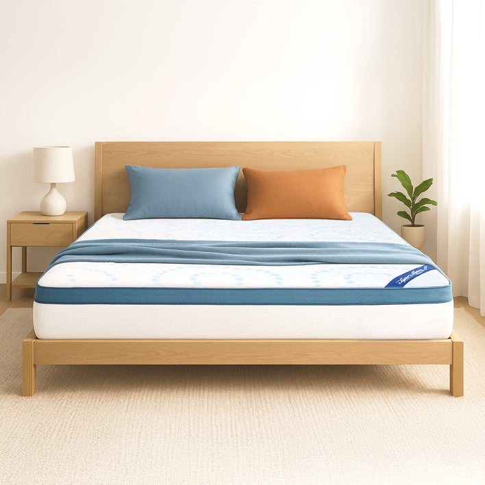 A simple, bright bedroom features a light wood bed with the Super Sleeper Pro [King] Straight 2 Sleep® Mattress Topper with Cooling Technology and FREE blue and orange pillows. A nightstand with a lamp and plant complete the hotel-like look.
