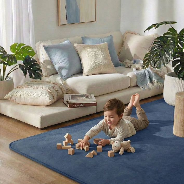 A young child plays with wooden blocks and a plush toy on the Super Sleeper Pro Plush Soft Touch Sensory Tatami Mat—a play mat that looks like a rug—in the living room, near a cream sofa with cushions and a potted plant on the wooden floor.