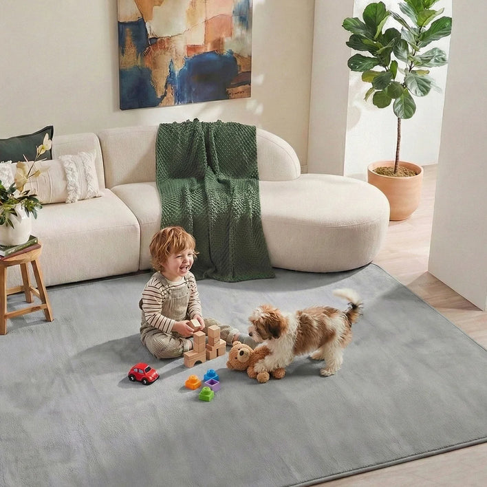 A young child plays with toys on the Super Sleeper Pro Plush Soft Touch Sensory Tatami Mat, which looks like a rug but works as a play mat, while a dog stands nearby holding a teddy bear near a beige sofa, green blanket, and potted plant.