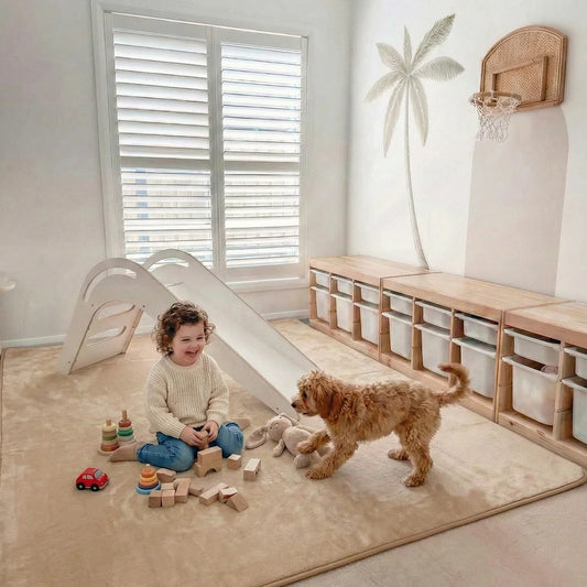 A young child sits, smiling at a small brown dog, on the Super Sleeper Pro Plush Soft Touch Sensory Tatami Mat—looks like a rug, works like a play mat. Wooden toys, blocks, and stuffed animals are scattered nearby in the vibrant playroom.