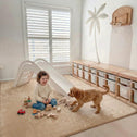 A young child sits, smiling at a small brown dog, on the Super Sleeper Pro Plush Soft Touch Sensory Tatami Mat—looks like a rug, works like a play mat. Wooden toys, blocks, and stuffed animals are scattered nearby in the vibrant playroom.