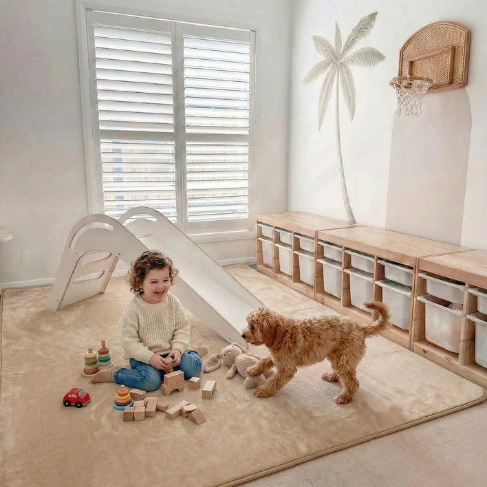A young child sits, smiling at a small brown dog, on the Super Sleeper Pro Plush Soft Touch Sensory Tatami Mat—looks like a rug, works like a play mat. Wooden toys, blocks, and stuffed animals are scattered nearby in the vibrant playroom.
