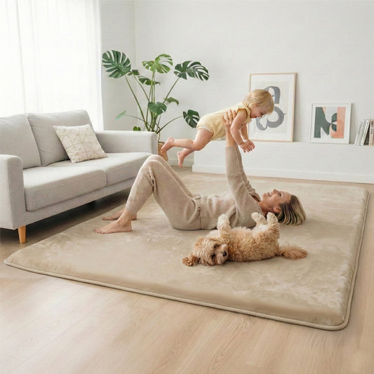 A woman relaxes on a Super Sleeper Pro Marshmellow™ Memory Foam Play Mat & Rug (35mm Plush Comfort) in a bright living room, lifting her toddler as their dog rests nearby. The space includes a gray sofa, plant, and framed art on light walls.