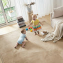 Two toddlers enjoy the Super Sleeper Pro Plush Soft Touch Sensory Tatami Mat in a bright living room—one stands in a yellow shirt and rainbow skirt, the other crawls in light blue. Toys, books, and a blanket add to the cozy rug-like play mat scene.