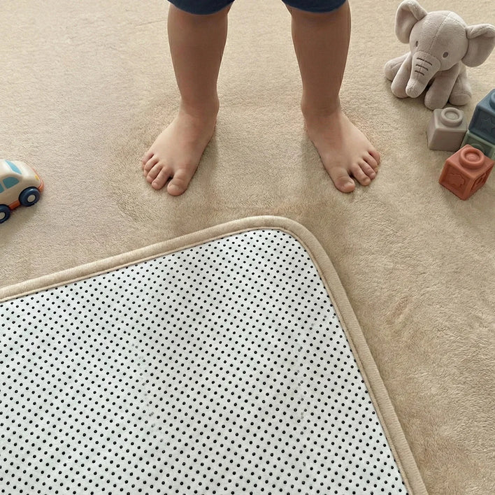 A child’s bare feet stand on the Super Sleeper Pro Plush Soft Touch Sensory Tatami Mat—looks like a rug, works like a play mat. Around are just their legs and feet, plus a toy elephant, colorful blocks, and a toy car.