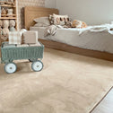 A neatly made bed with checkered bedding and stuffed animals sits against a scalloped wall; a green toy wagon with gift boxes is on the Super Sleeper Pro Plush Soft Touch Sensory Tatami Mat, which looks like a rug but works as a play mat in this tidy, softly-lit room.