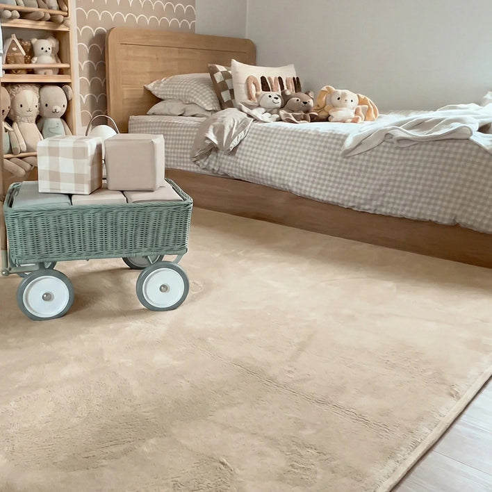 A neatly made bed with checkered bedding and stuffed animals sits against a scalloped wall; a green toy wagon with gift boxes is on the Super Sleeper Pro Plush Soft Touch Sensory Tatami Mat, which looks like a rug but works as a play mat in this tidy, softly-lit room.