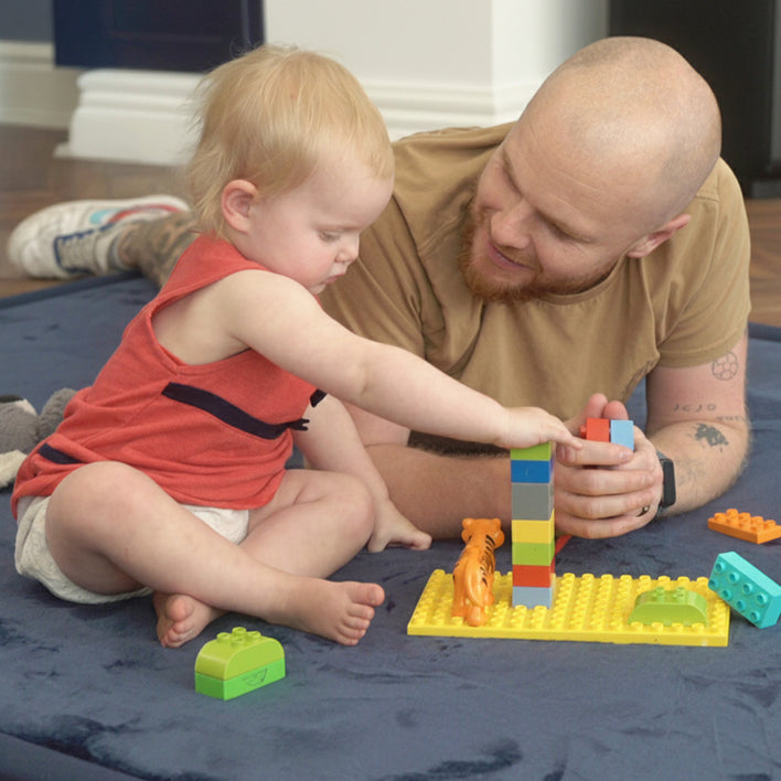 Toddler playing with building blocks on a soft tatami floor mat while an adult watches and supports playtime.