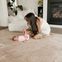 A woman with long dark hair and tattoos kneels on the Plush Soft Touch Sensory Tatami Mat by Super Sleeper Pro, gently holding a baby’s feet. In the background are a white chair, plants, and a fireplace.