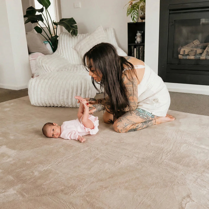 A woman with long dark hair and tattoos kneels on the Plush Soft Touch Sensory Tatami Mat by Super Sleeper Pro, gently holding a baby’s feet. In the background are a white chair, plants, and a fireplace.