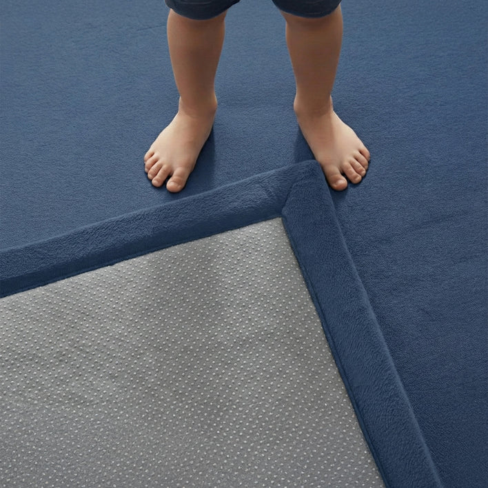 Close-up of a child standing on a blue floor mat with the corner folded to reveal a grey anti-slip underside featuring white dotted grip texture.