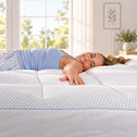 A smiling woman in a light blue top lies on her side on a white quilted mattress topper in a bright, sunlit bedroom.