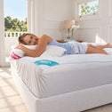 A woman lies on a white bed with a white pillow and a white mattress protector featuring a blue and pink Frosty label, in a bright, sunlit bedroom.