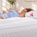 A woman lies on a white quilted mattress in a bright bedroom, with a pink pillow featuring the brand name 'SLEEP WELL ANY' visible.