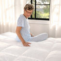 An older woman with short curly gray hair and glasses sits cross-legged on a white quilted comforter in a bright bedroom. She is wearing a light blue pajama top with small white motifs and matching light blue striped pajama pants.