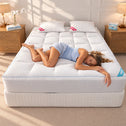 A woman lies on her side on a white quilted mattress on a mattress base in a bright, modern bedroom. The mattress has a white quilted top and a white ribbed base, with two white pillows and wooden bedside tables with lamps on either side.